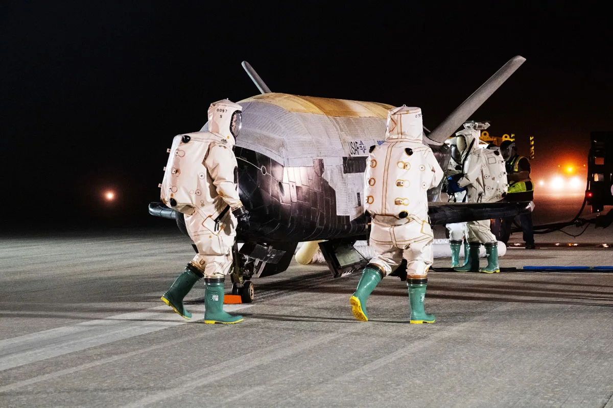 x-37b-and-people People in hazmat-like suits approach the X-37B on the runway.